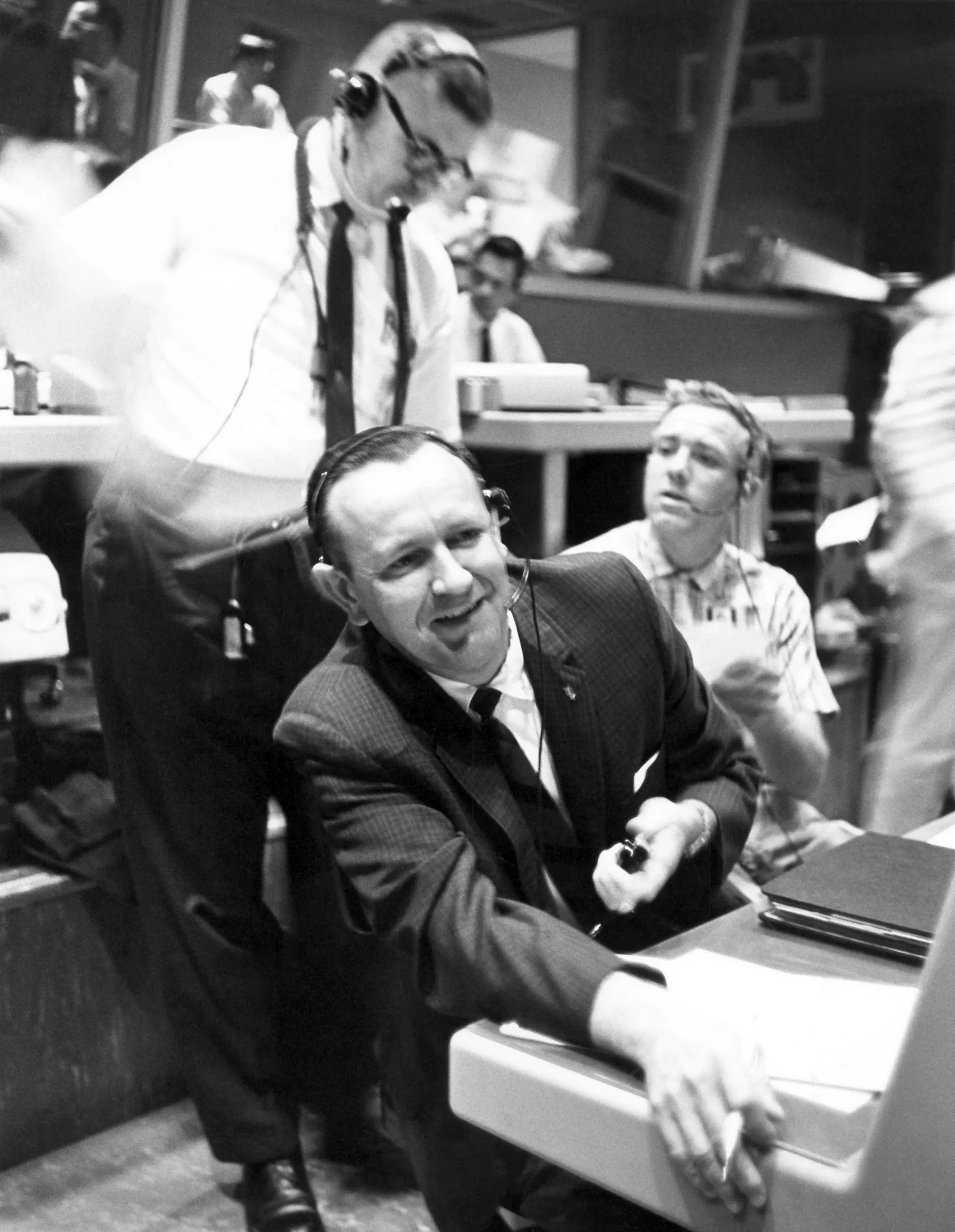 Christopher Kraft, in shirtsleeves and a headset, leans on his console while colleagues stand behind him.