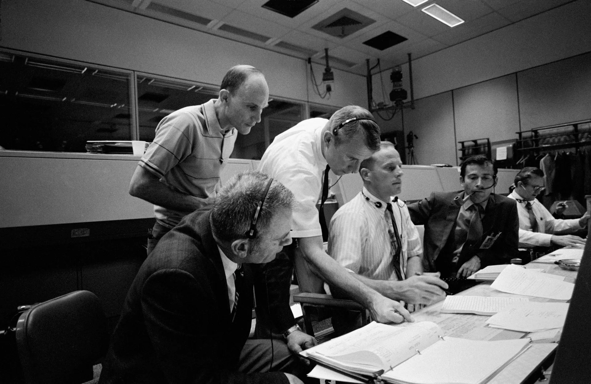 Five astronauts in shirtsleeves huddle over flight plans at a console in Mission Control.