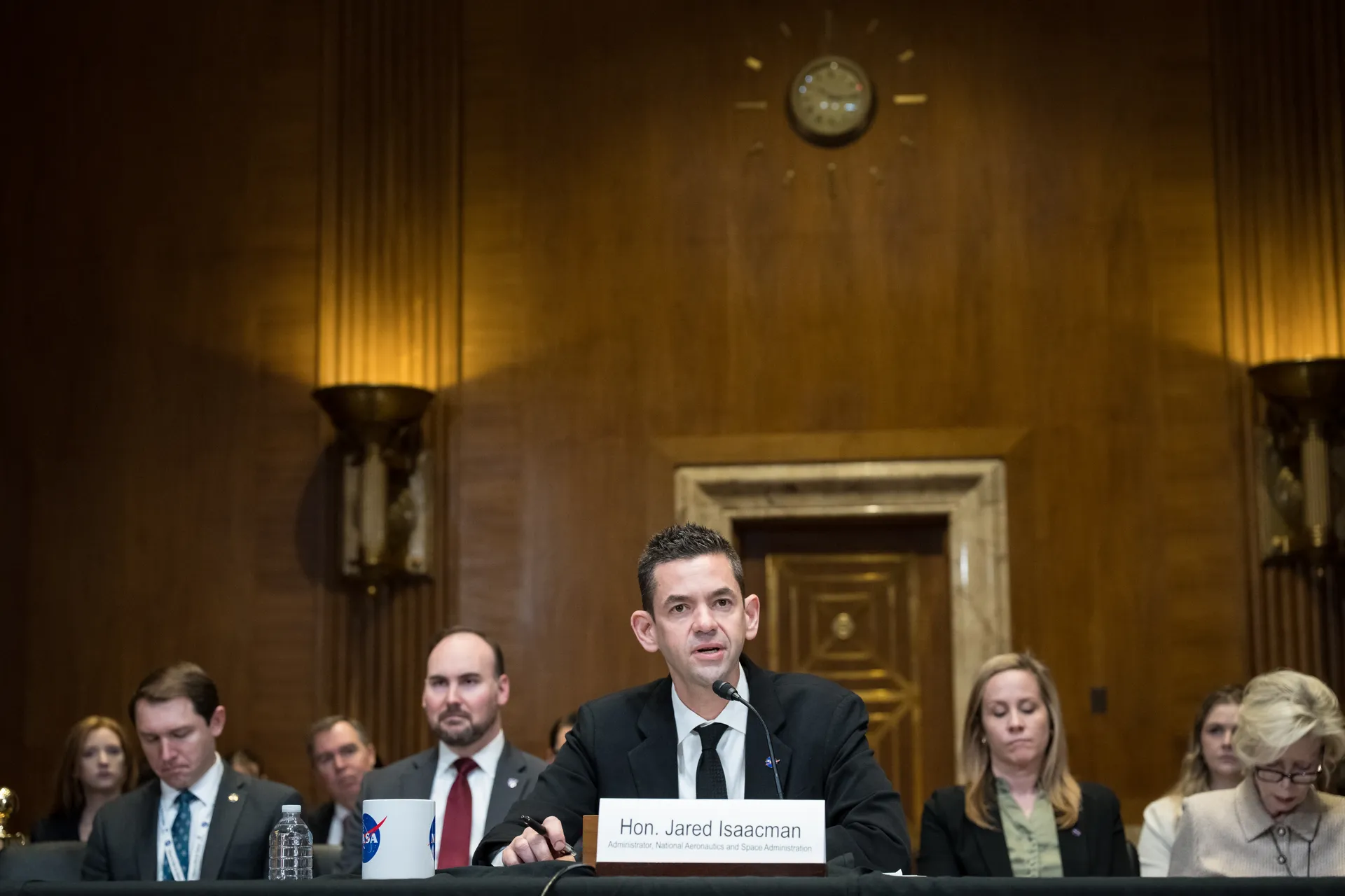 NASA Administrator Jared Isaacman testifies at a witness table in a wood-paneled Senate hearing room, staff seated on either side.