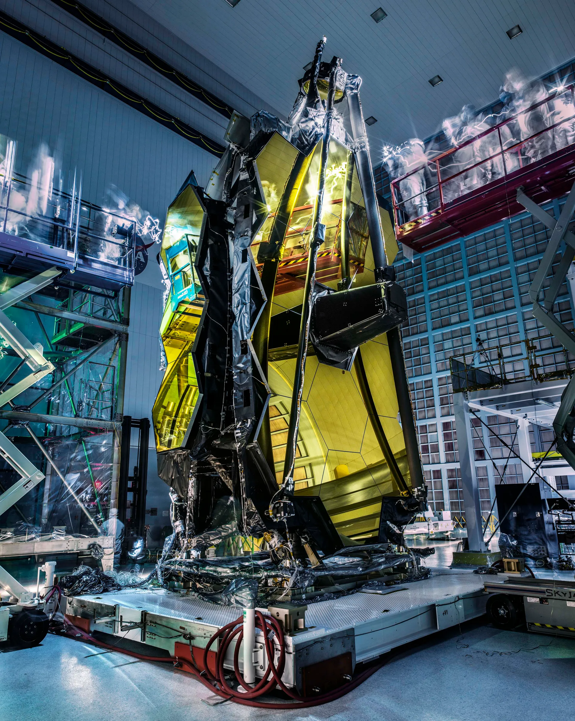 Technicians appear as ghostly blurs beside the folded James Webb Space Telescope and its gold mirrors in a dark clean room.