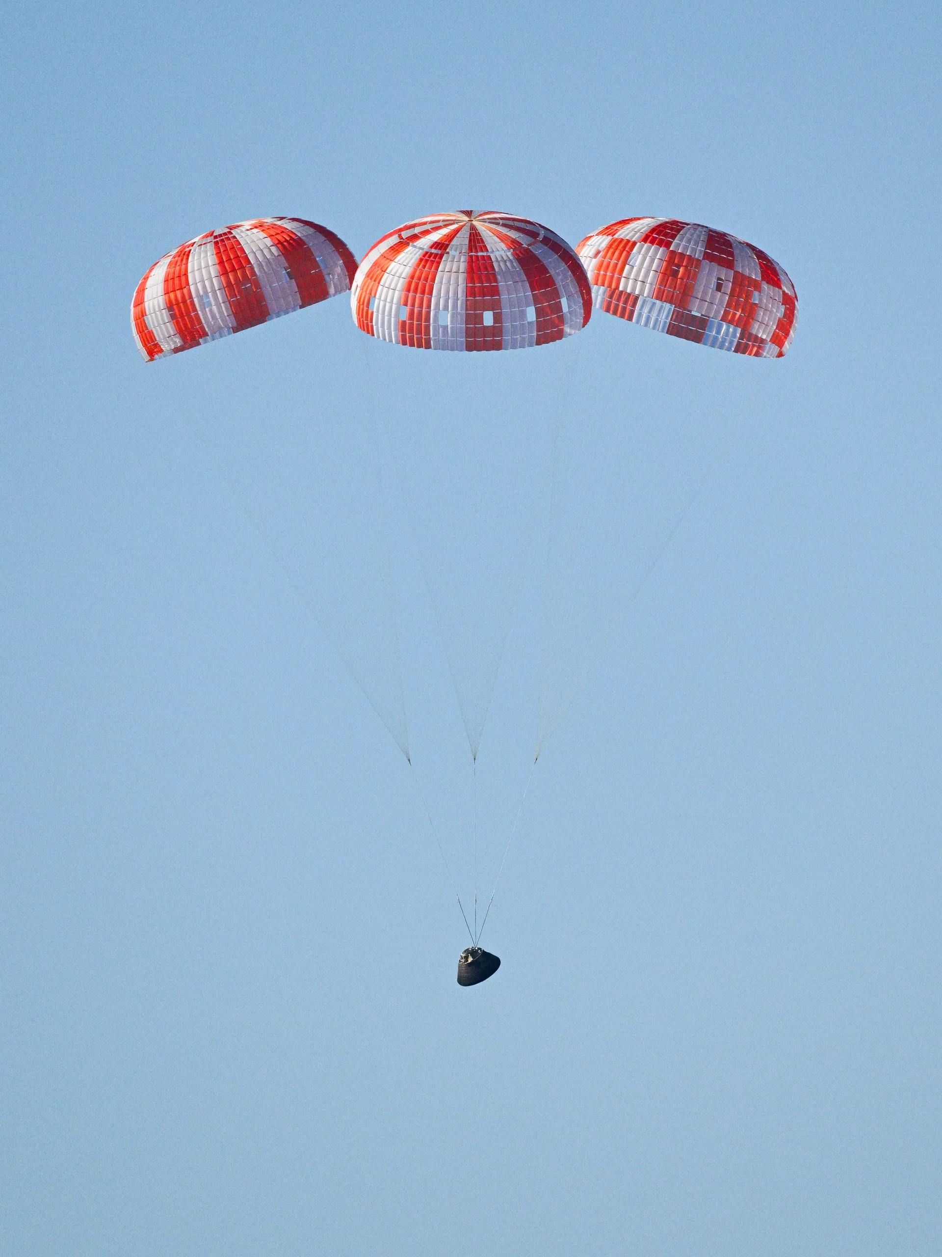 Three red-and-white striped parachutes lower the small Orion capsule toward the Pacific under a clear blue sky.