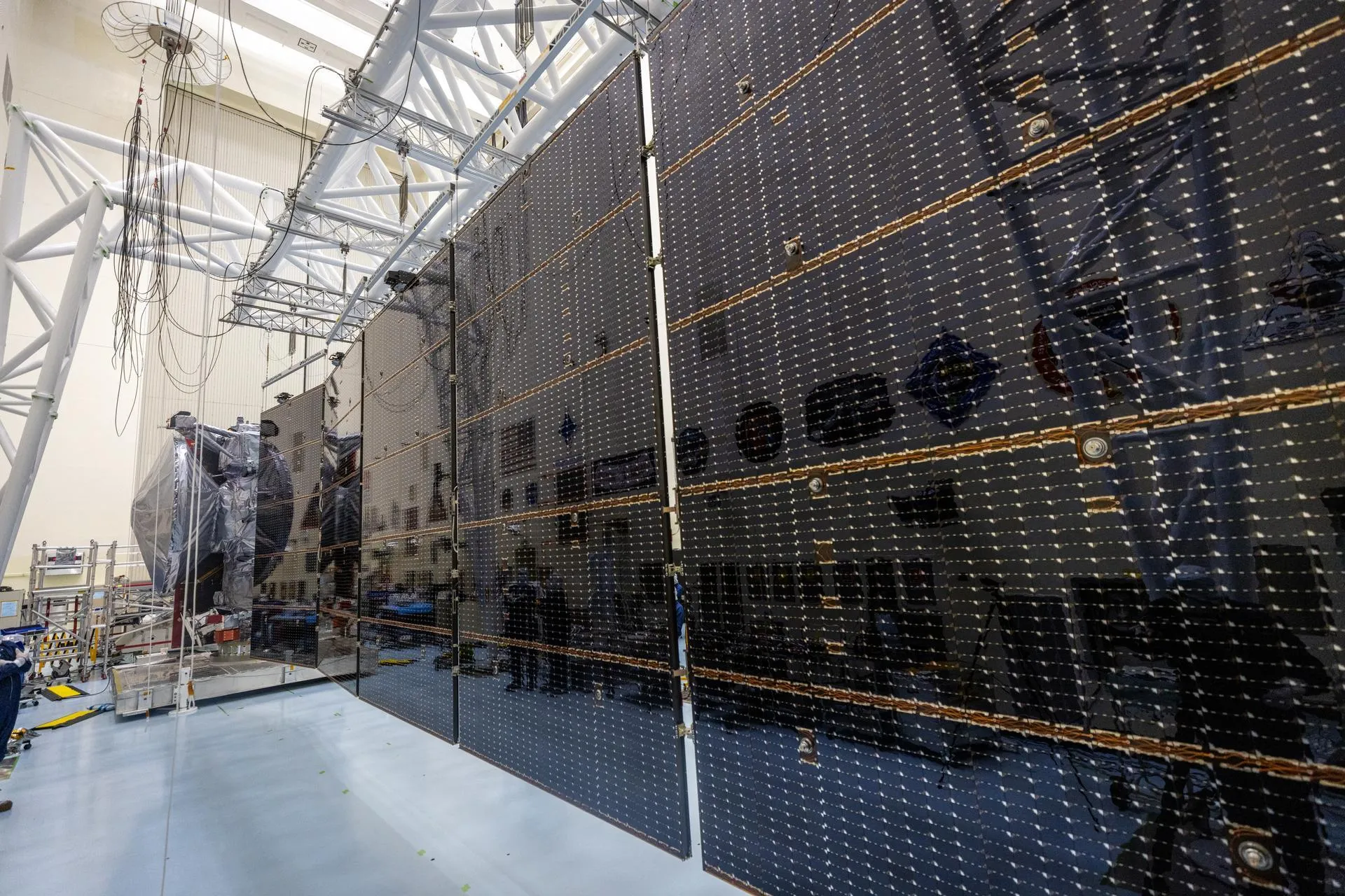 A long wall of dark solar cells stretches away across a high clean room, with the spacecraft body small at the far end.