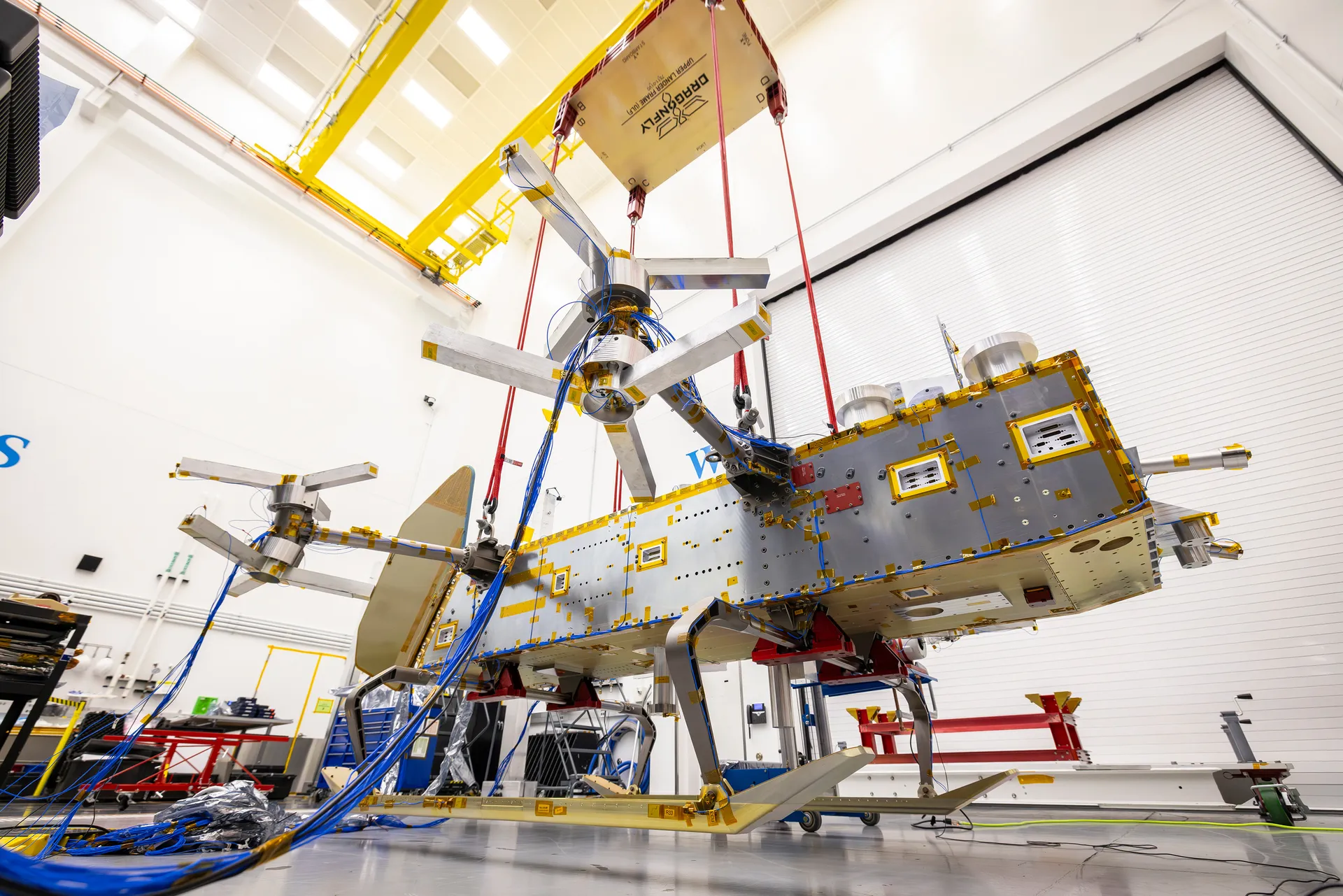 A long silver and gold box-shaped frame with rotor arms hangs from bungee cords inches above a clean-room floor.