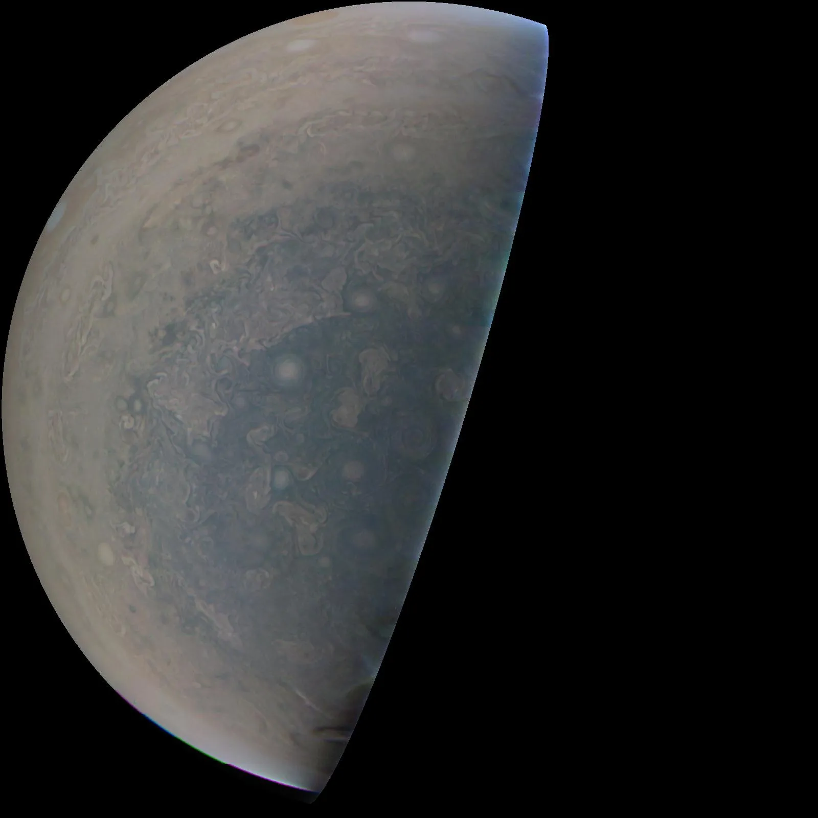Half of Jupiter’s disk seen from below the south pole: a bluish-gray field of swirling oval storms fading to tan bands at the edge, against black space.