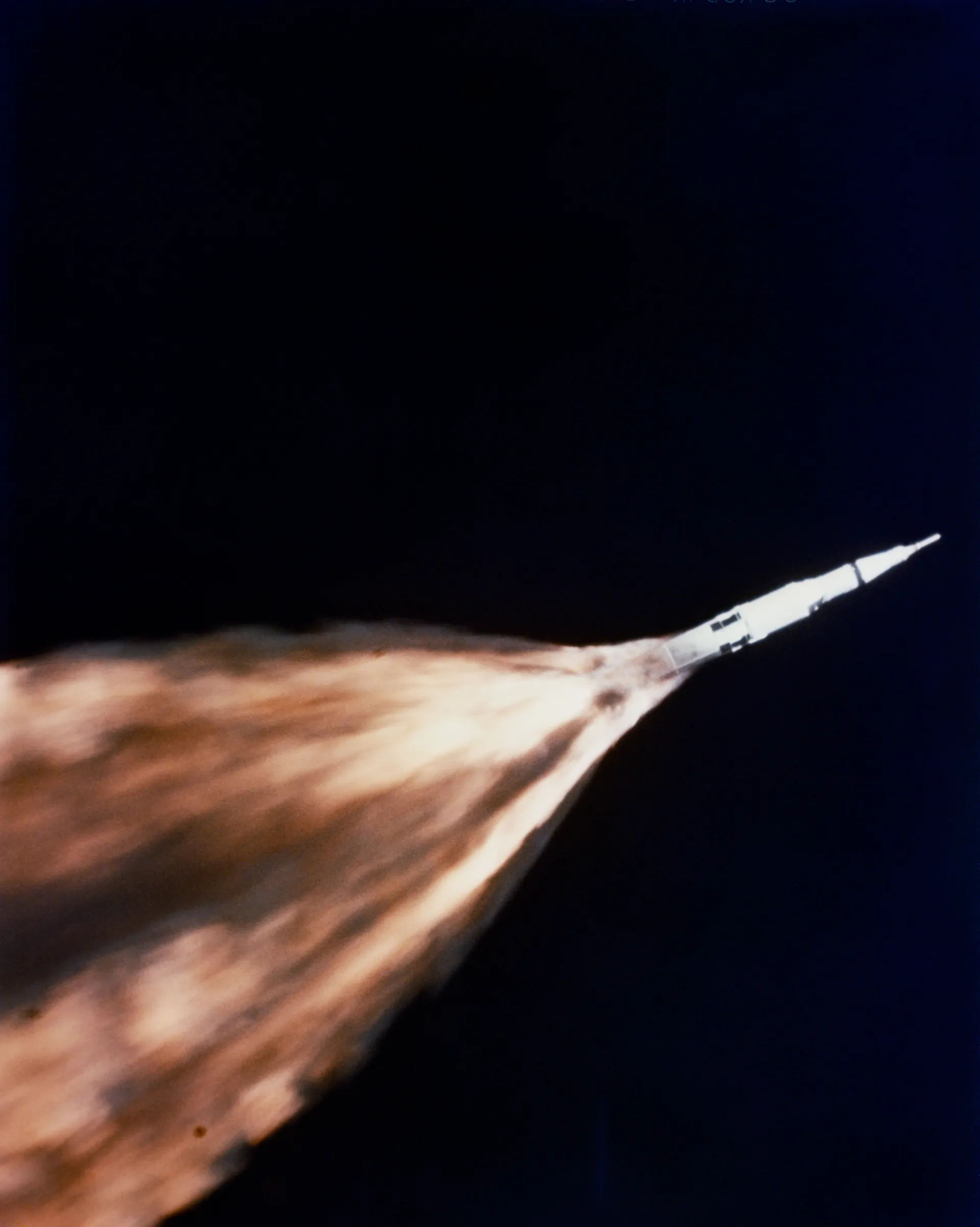 Seen from a chase plane against a dark blue sky, a Saturn V climbs to the upper right trailing an enormous orange plume of flame.