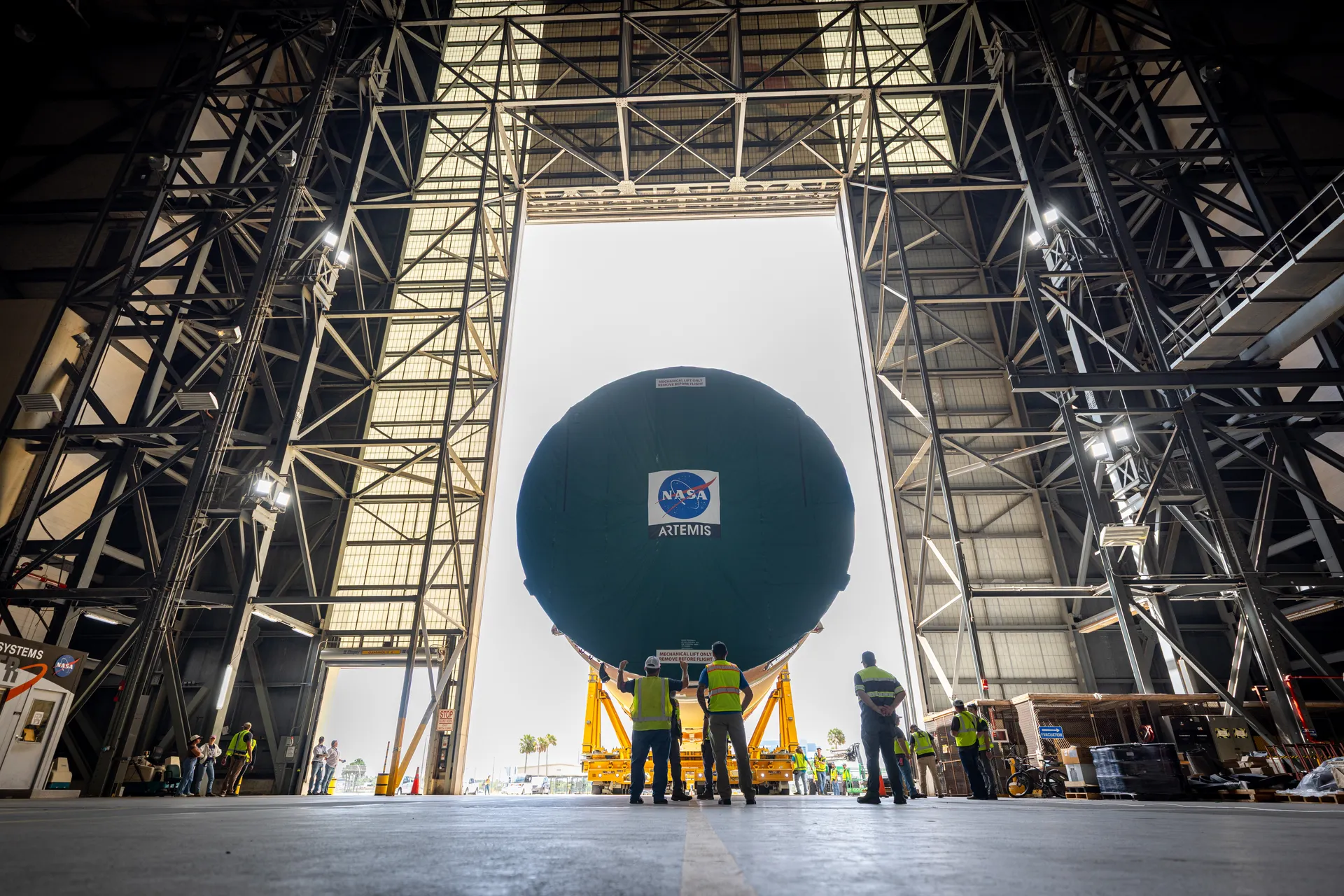 Inside the Vehicle Assembly Building, workers stand beneath the round end of a large core stage section that fills the tall doorway.