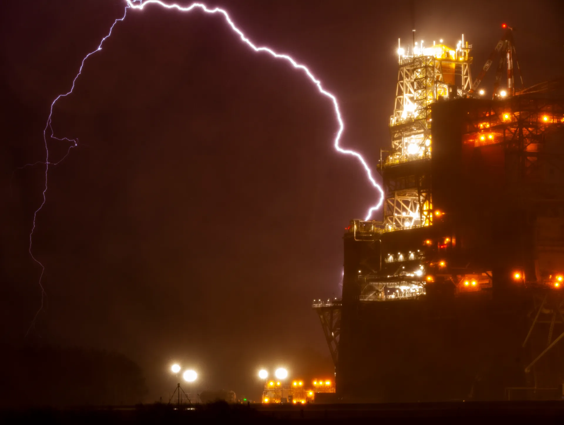 At night, a bolt of lightning arcs across the sky beside a brightly lit steel test stand holding the SLS core stage.