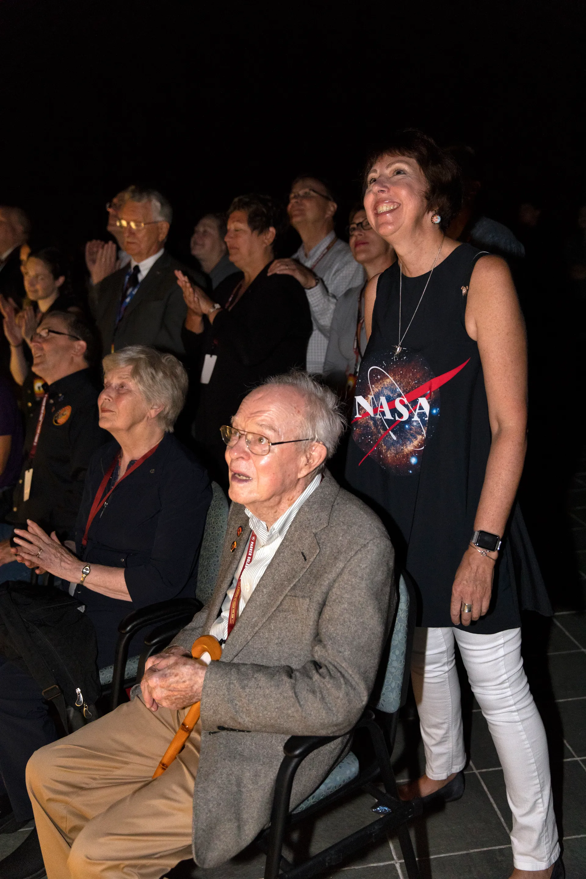 An elderly man in a tweed jacket sits in front of a crowd at night, looking up and to the right; the people around him are smiling and pointing upward.