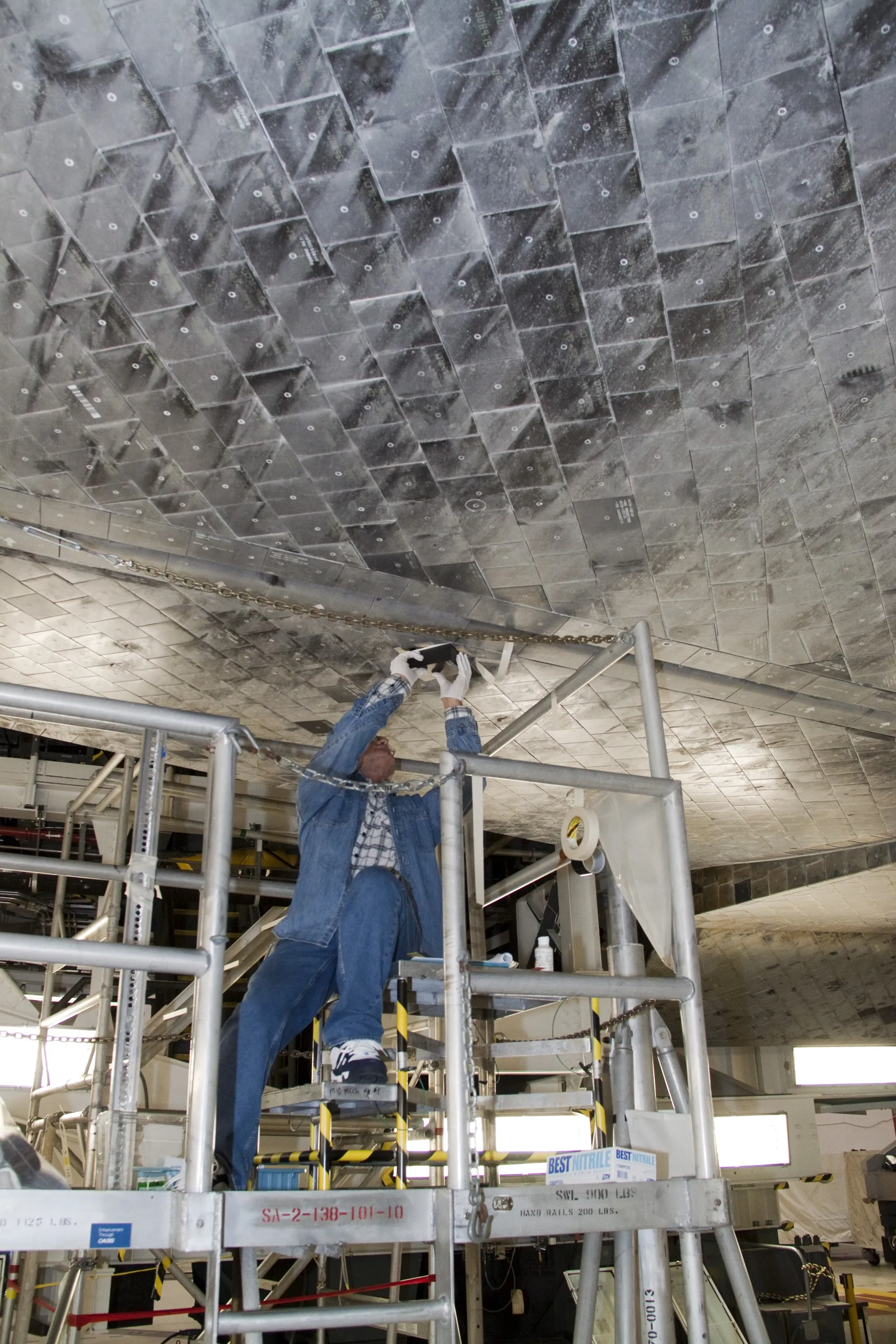 A technician on a work stand replaces a black thermal tile on the underside of Atlantis, surrounded by rows of tiles.