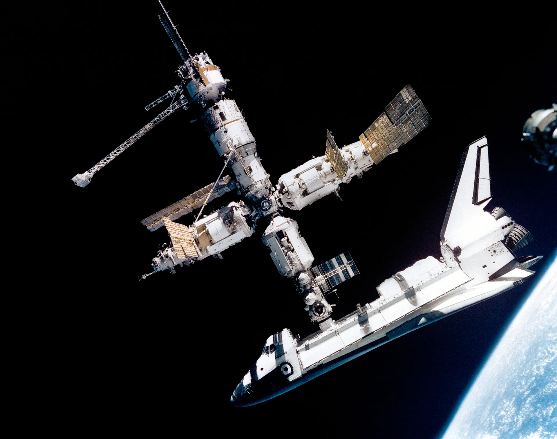 Atlantis docked to the Russian Mir station against black space, with Earth’s edge in the corner.