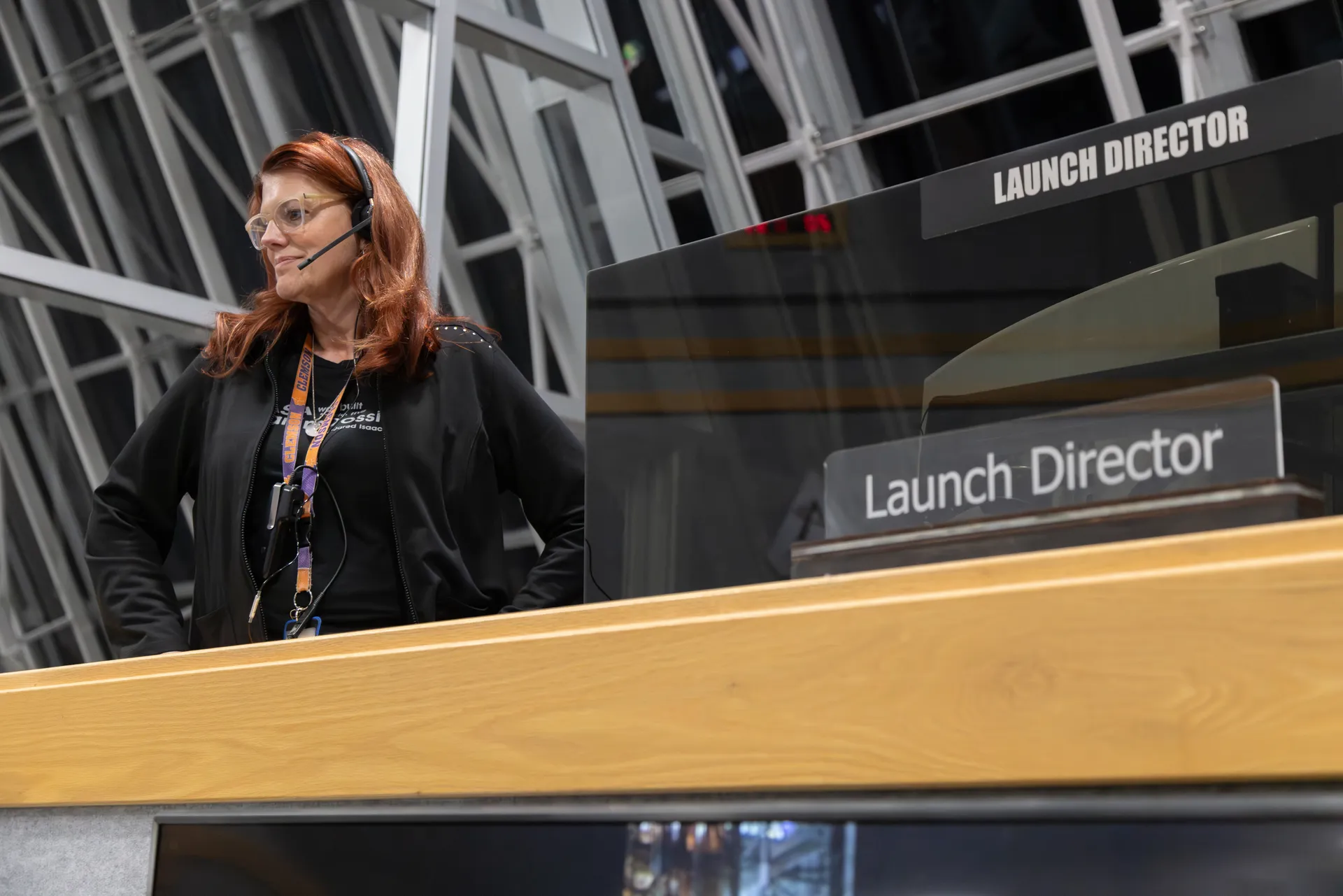 A launch director in a headset stands at a raised console labeled Launch Director, with tall windows behind her in a control room.