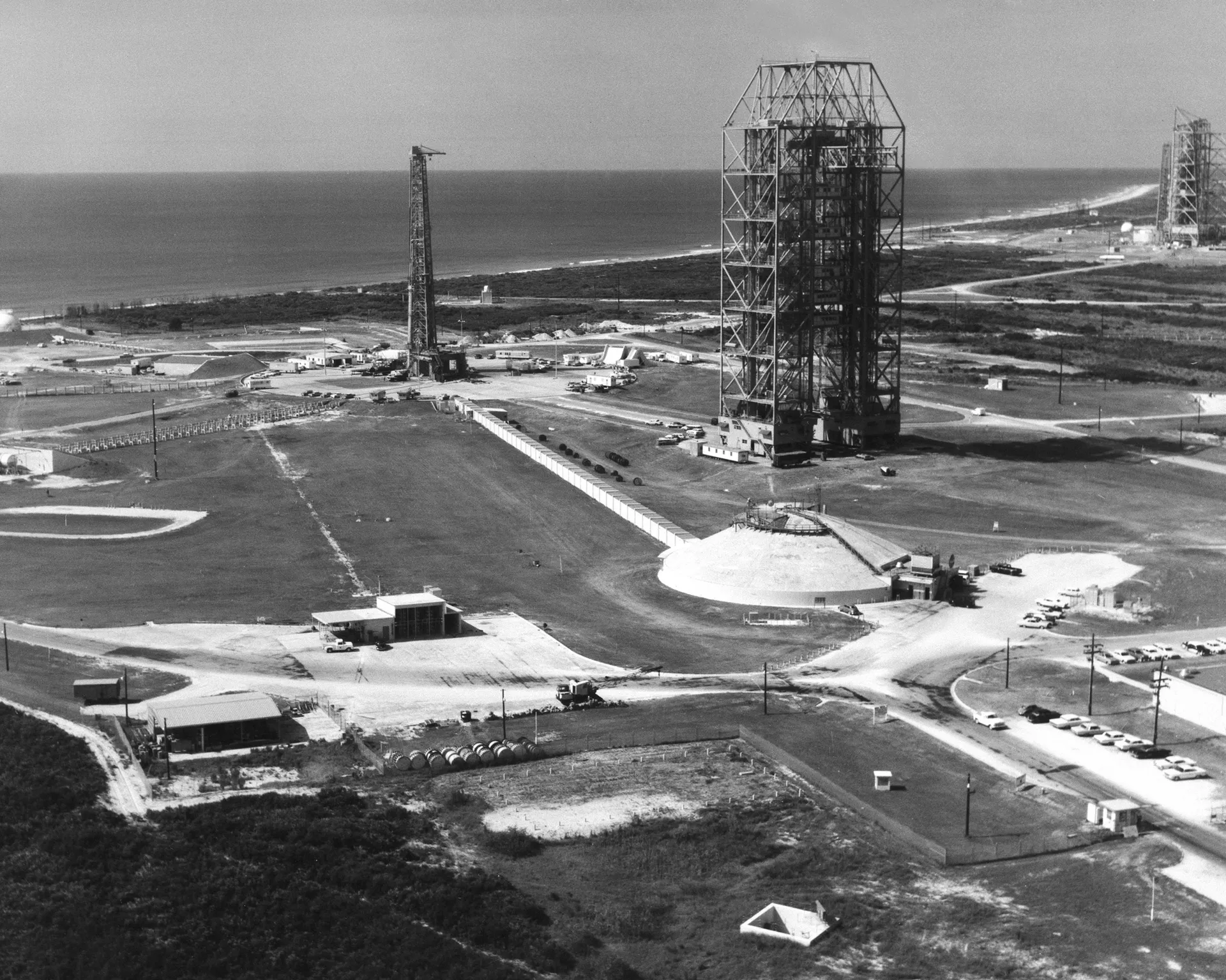 Black-and-white aerial view of a launch complex on a sandy coast: a tall service tower, a domed blockhouse and a launch pedestal, with the ocean beyond.