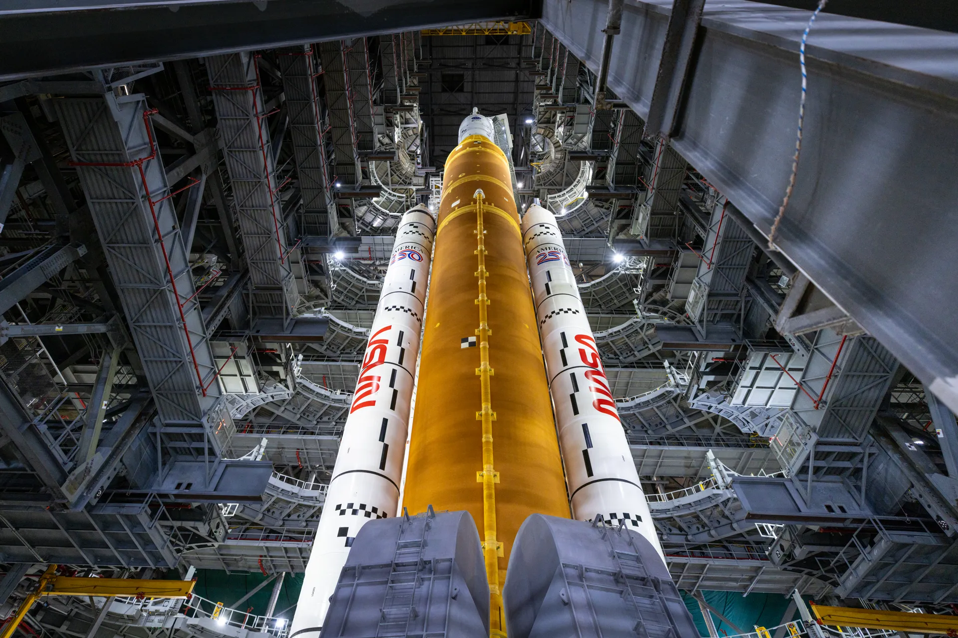 Looking up inside the Vehicle Assembly Building at the orange SLS core stage and two white boosters, surrounded by the steel framework of retracted work platforms.