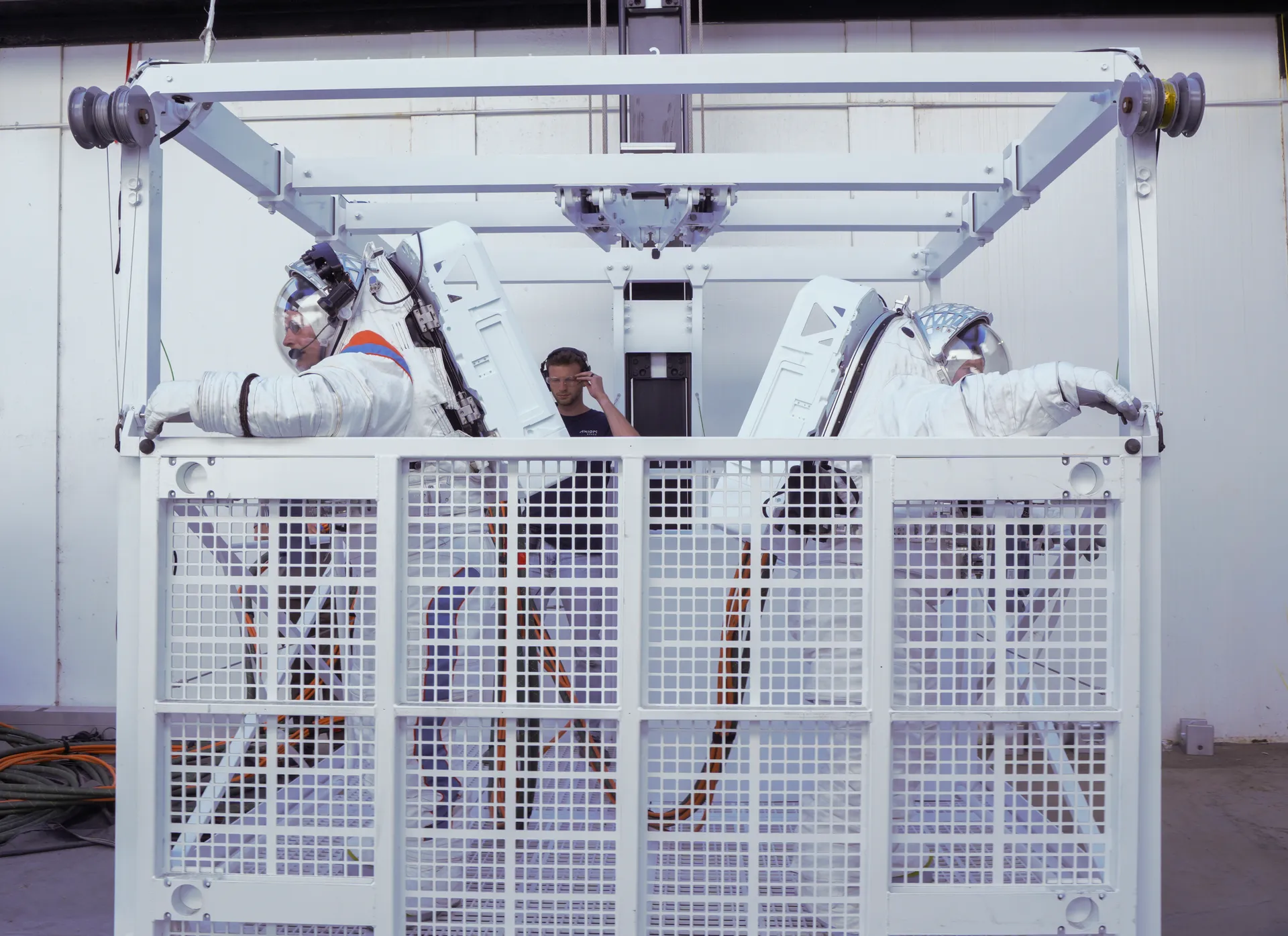 Two suited astronauts stand in a white mesh elevator basket hanging from a steel frame, in a test facility.