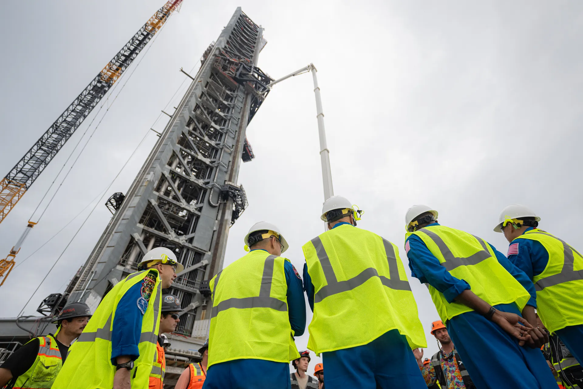 Astronauts in blue flight suits and safety vests look up at a tall steel launch tower under construction.