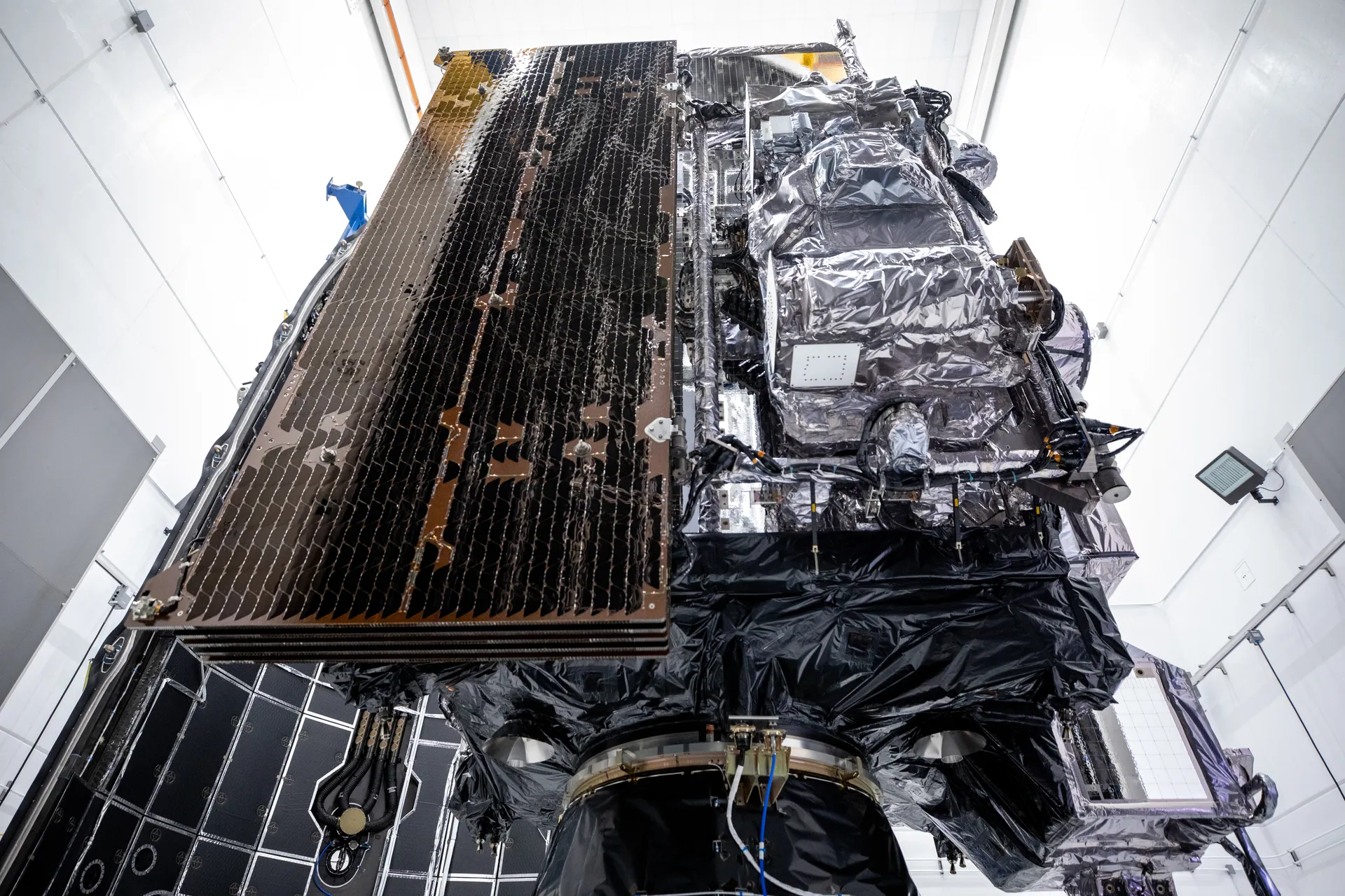 GOES-U seen from below in a white clean room: a dark folded solar array on one side, instruments wrapped in silver insulation on the other.